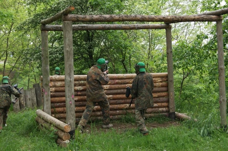 Grupa osób w kombinezonach moro, chowająca się za drewnianą ścianką